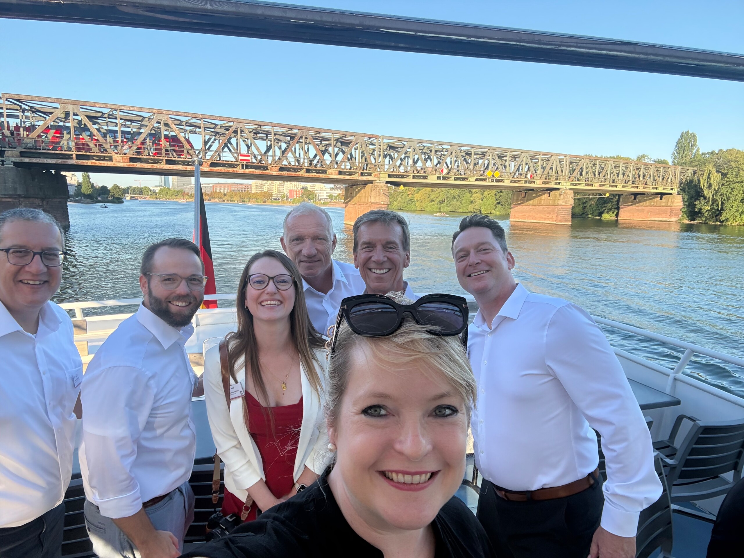 Sieben Teilnehmende, unter ihnen Rechtsanwältin Simone Krämer und Rechtsanwalt Jan Lehniger, machen auf dem Schiffsdeck ein gemeinsames Selfie vor einer Mainbrücke in Frankfurt.